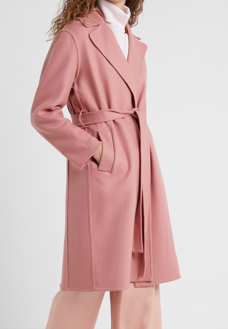 Růžový kabát z vlněné směsi s páskem v pase, klopovým límcem a bočními kapsami. Hladká textura a na míru šitý střih, stylizovaný s pastelovými kalhotami.