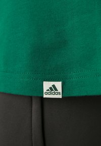 Camiseta de algodón verde con un cuello acanalado y una etiqueta de logo de Adidas en la esquina inferior, combinada con una tela negra abajo.
