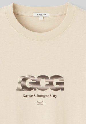 T-shirt à manches courtes couleur crème affichant un logo "GCG" rayé marron et le texte "Game Changer Guy", avec un col ras du cou côtelé. Taille indiquée : L.