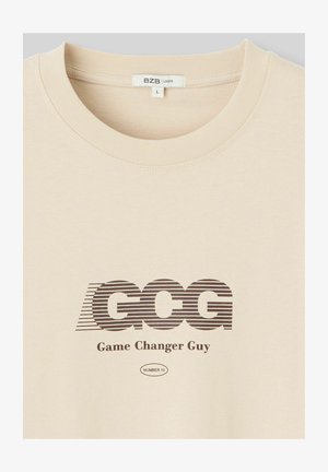 T-shirt à manches courtes couleur crème affichant un logo "GCG" rayé marron et le texte "Game Changer Guy", avec un col ras du cou côtelé. Taille indiquée : L.