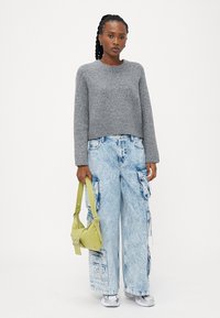 Maglione grigio corto, ampi pantaloni cargo in denim azzurro chiaro con tasche, e una piccola borsa verde. Texture liscia, vestibilità rilassata.