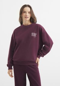 Sudadera morada con cuello redondo y mangas largas, que presenta un bordado blanco de "Chelsea" en la zona del pecho izquierdo. Textura de tela suave.