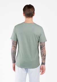 Mężczyzna z krótkimi brązowymi włosami, wytatuowanymi ramionami, noszący zieloną koszulkę i białe szorty, stojący tyłem na białym tle.