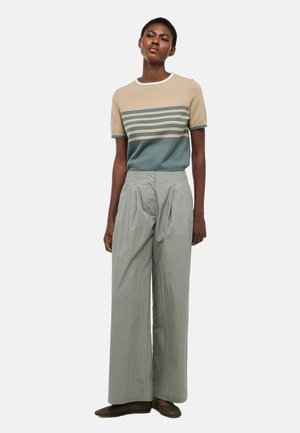 Femme debout portant un haut à manches courtes beige rayé de vert, un pantalon ample gris clair taille haute et des chaussures sombres sans lacets.