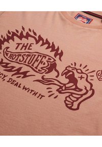 Camiseta de algodón rosa claro con diseño gráfico en color marrón que presenta una mano en llamas y una serpiente, el texto dice "LO BUENO, PICANTE, AFRÓNTALO."