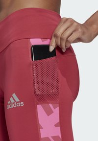 Rote Sportleggings mit einer Netzeltasche, die für die Aufbewahrung des Handys ausgelegt ist. Sie verfügen über ein pinkes geometrisches Muster und ein silbernes adidas-Logo.