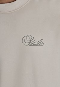 Beige katoenen T-shirt met een ronde hals, met aan de voorkant een subtiel grijs merkembleem "Sitsilk". Soepele textuur, minimalistisch ontwerp.