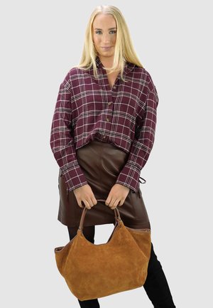 Blonde vrouw die een bordeauxrood geruit overhemd en een bruine leren rok draagt, steekt een grote beige suède handtas vast, staand tegen een grijze achtergrond.