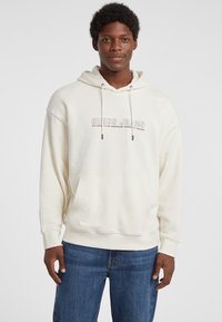 Sudadera blanca hecha de algodón suave, con un bolsillo frontal, capucha con cordón y texto impreso "GUESS JEANS" en colores contrastantes.
