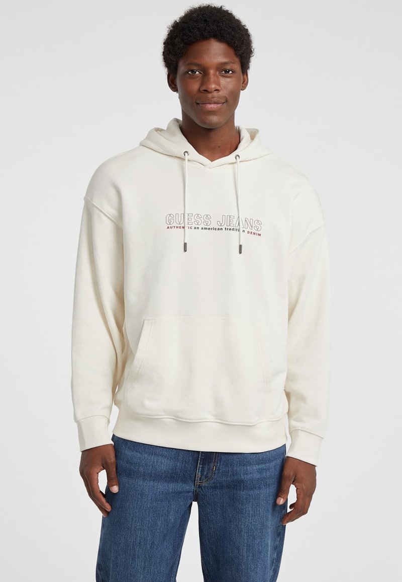 Sudadera blanca hecha de algodón suave, con un bolsillo frontal, capucha con cordón y texto impreso "GUESS JEANS" en colores contrastantes.