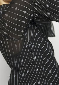 Top in tessuto nero trasparente con strisce orizzontali bianche e piccoli motivi circolari. Le maniche sono a sbuffo, creando texture e dimensione.