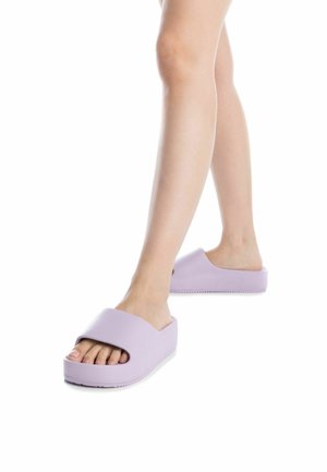 Zapatillas moradas de tipo slip-on con una banda ancha y punta abierta. Fabricadas con material suave y suela gruesa y texturizada. Diseño minimalista.
