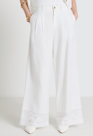 Weit geschnittene weiße Hosen mit Lochspitzenbesatz an den Säumen, getragen mit weißen spitzen Schuhen und goldenen Armreifen an den Händen in den Taschen.