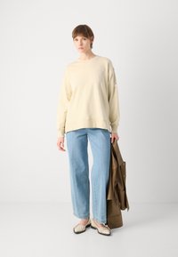Felpa color crema dal taglio morbido, abbinata a jeans a gamba larga di un blu chiaro. Il modello tiene in mano una giacca marrone e indossa scarpe beige.