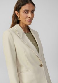 Blazer beige avec un col cranté, fermeture à un bouton, et poches latérales, associé à une chemise verte à motifs en points.
