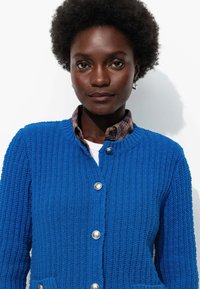 Cardigan bleu texturé avec un design côtelé, comportant des boutons argentés et un col de chemise à carreaux replié en dessous.
