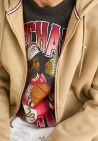 Beżowa bluza z kapturem z czerwonymi i białymi akcentami na czarnym T-shircie z kolorową grafiką koszykówki, przedstawiającą zawodnika i piłkę do koszykówki.