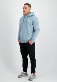 Sudadera azul claro con un bolsillo canguro, capucha con cuerda y un pequeño logo. Combinada con pantalones cargo negros y zapatillas grises.