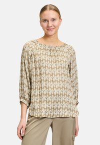 Top beige con un motivo geometrico in verde tenue e bianco. Presenta una scollatura ampia e maniche a tre quarti con polsini arricciati.