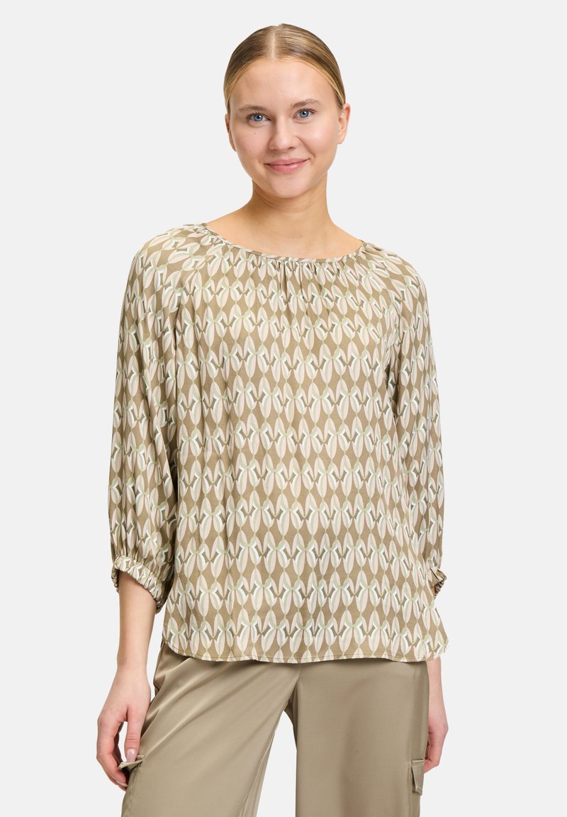 Top beige con un motivo geometrico in verde tenue e bianco. Presenta una scollatura ampia e maniche a tre quarti con polsini arricciati.