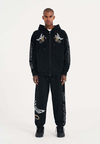 Schwarze Zip-Hoodie und passende Hose, mit gestickten Flügel- und Schlangendesigns, "ED HARDY"-Text, dickem Stoff und lockerer Passform.