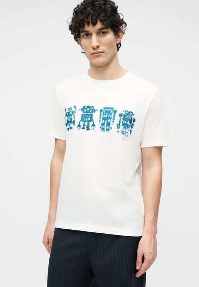 Άνδρας με σγουρά σκούρα μαλλιά, ντυμένος με άσπρο μπλουζάκι που έχει εκτυπωμένα τέσσερα μπλε στυλιζαρισμένα ρομπότ, συνδυασμένο με σκούρο παντελόνι με ρίγες.