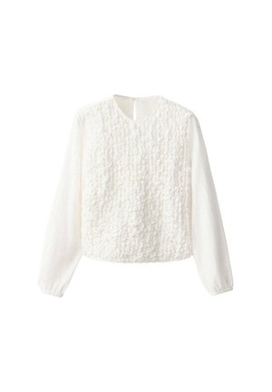 Joven mujer con top blanco texturizado de manga larga, vaqueros azules holgados y zapatos burdeos, llevando un bolso pequeño de hombro con una correa ancha.