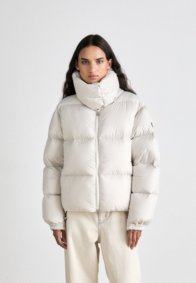 Puha lahka jakna svetlo sive barve z visokim ovratnikom, zadrgo in elastičnimi manšeti. Ima škatlasto obliko in gladko teksturo.