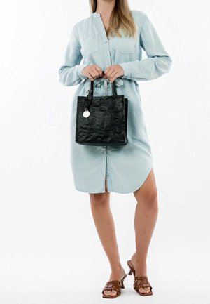 Femme en robe boutonnée bleu clair tenant un sac à main noir texturé, portant des sandales à talons marron sur fond blanc.