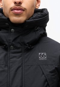 Homme portant une veste noire 66° North avec col montant et détails de boutons, visage partiellement visible du nez vers le bas.