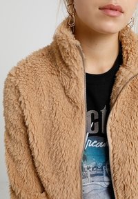 Beżowa kurtka na zamek z imitacji futra o teksturowanej, puszystej powierzchni, z kołnierzem stójką, noszona na czarnej koszulce z nadrukiem.