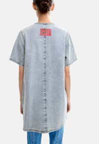 Jasnoniebieska, oversize sukienka koszulowa z denimu, z krótkimi rękawami, centralnym szwem oraz czerwoną naszywką z logo na plecach, charakteryzująca się miękką teksturą.