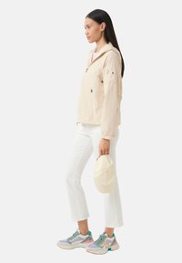 Femme en veste à capuche beige, pantalon blanc court, baskets colorées, tenant une casquette beige, se tenant de profil sur un fond blanc uni.
