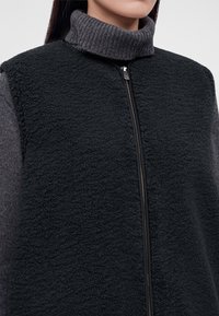 Femme portant un gilet polaire noir zippé par-dessus un pull col roulé côtelé gris foncé, cadré du cou au milieu du torse.