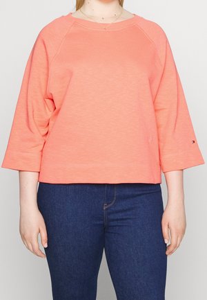 Kvinde iført koralrosa top med trekvartærmer og mørkeblå jeans, stående mod en ensfarvet lys baggrund.