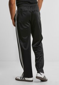 Black athletic pants with white side stripes, elastic waistband, and two back pockets. Material appears smooth and slightly shiny.