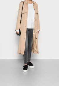 Beige Trenchcoat mit taillenlangem Schnitt, Knopfleiste und Schulterklappen, kombiniert mit einem weißen Hemd, grauen Jeans und schwarzen Sneakers.
