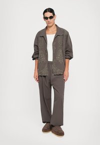 Brun mønstrede oversized jakke med lynlås, hvid tanktop, matchende brune bukser og brune slip-on sko. Sorte solbriller.