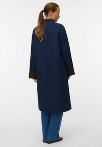 Lungo cappotto blu navy con dettagli marroni ai polsini, caratterizzato da un design semplice e una texture liscia. La vista posteriore mostra un taglio dritto fino a metà polpaccio.