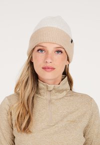 Pletena beanie u krem i bež boji s rebrastom teksturom, koja ima preklopeni rub. Nosena s bež majicom s visokim ovratnikom i zatvaračem.
