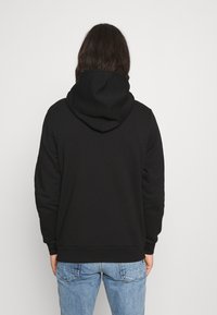 Černá mikina s kapucí na šňůrku, žebrovanými manžetami a spodním lemem, vyrobena z měkké látky, zobrazená zezadu, v kombinaci se světle modrými džíny.