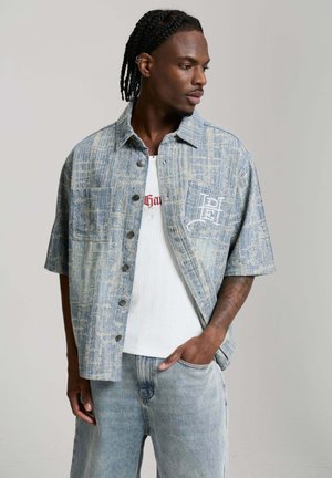 Mann mit geflochtenem Haar, der ein gemustertes kurzärmeliges Hemd über einem weißen gerippten Top und hellen Jeans trägt, Hand in der Tasche, schaut nach rechts.