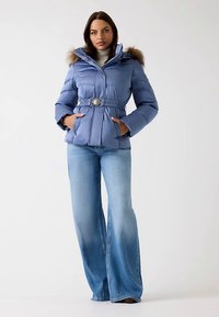 Gaiši zila pūkaina jaka ar kažokādas oderētu kapuci, jostasvietu ar jostu un sānu kabatām. Kombinēta ar izplestām, izbalinātām zilām džinsiem.