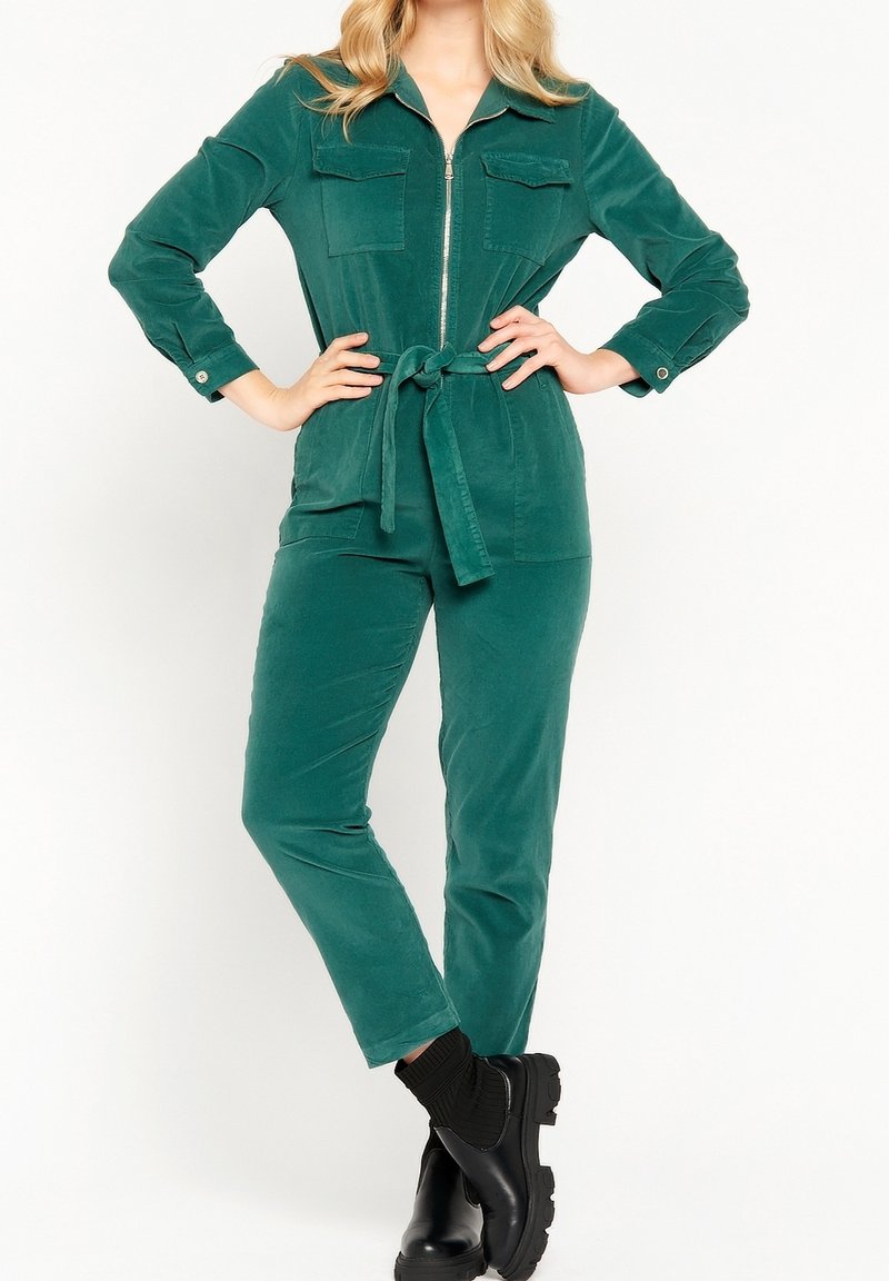 Femme debout en combinaison verte à fermeture éclair, ceinturée, avec poches avant, portant des bottines noires épaisses, sur fond blanc.