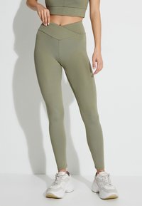 Leggings verde oliva con una cintura alta cruzada, hechos de un material suave y elástico. Combinados con zapatillas blancas gruesas.