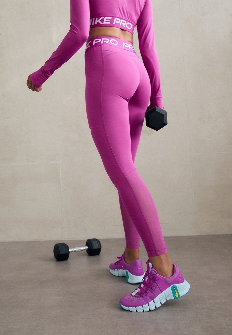 Różowe legginsy sportowe i long-sleeve top, oba z logo „Nike Pro”. Obuwie ma różowe i akwamarynowe akcenty z teksturowaną podeszwą. Obok znajduje się regulowany hantle.