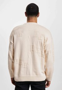Maglione beige a maglia con scollo rotondo, dettagli del logo testurizzati e polsini a costine. Fit rilassato con un design a schiena semplice.