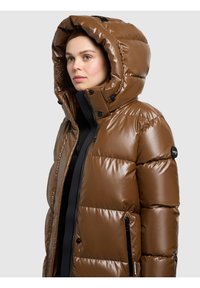 Jauna moteris dėvi blizgantį rudą pūstinę žieminę striukę su kapišonu, stovinti priešais paprastą šviesiai pilką foną, žiūrinti į šoną.