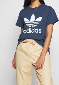 Námořnický bavlněný tričko s velkým bílým logem Adidas a designem tří pruhů. V kombinaci s béžovými kalhotami na šňůrku. Hladká textura.