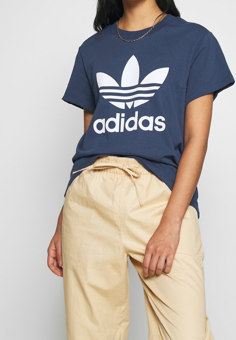 Námořnický bavlněný tričko s velkým bílým logem Adidas a designem tří pruhů. V kombinaci s béžovými kalhotami na šňůrku. Hladká textura.